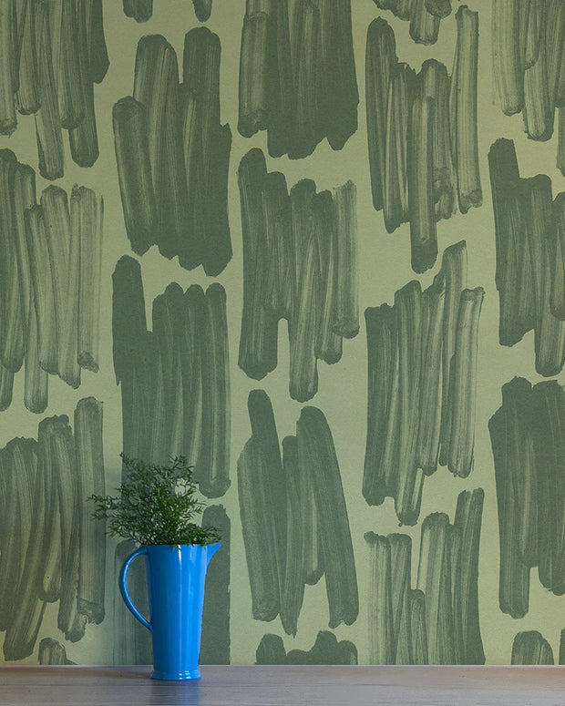 A section of wallpaper with an abstract geometric pattern in green shades, trimmed to show the digitally printed nonwoven texture, with a blue cup and a green plant to the side.