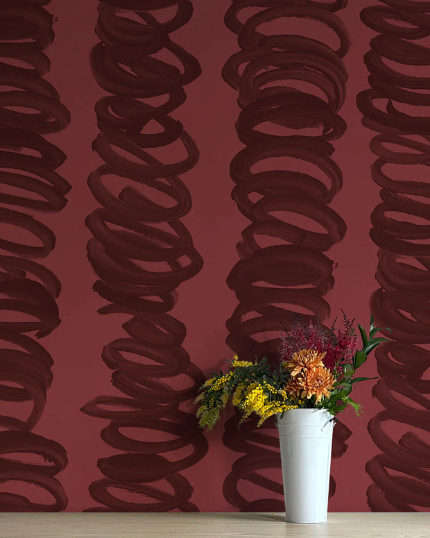 A roll of wallpaper with an abstract, contemporary design featuring a chocolate covered strawberry motif, positioned against a red background with a vase of dried flowers in the foreground.