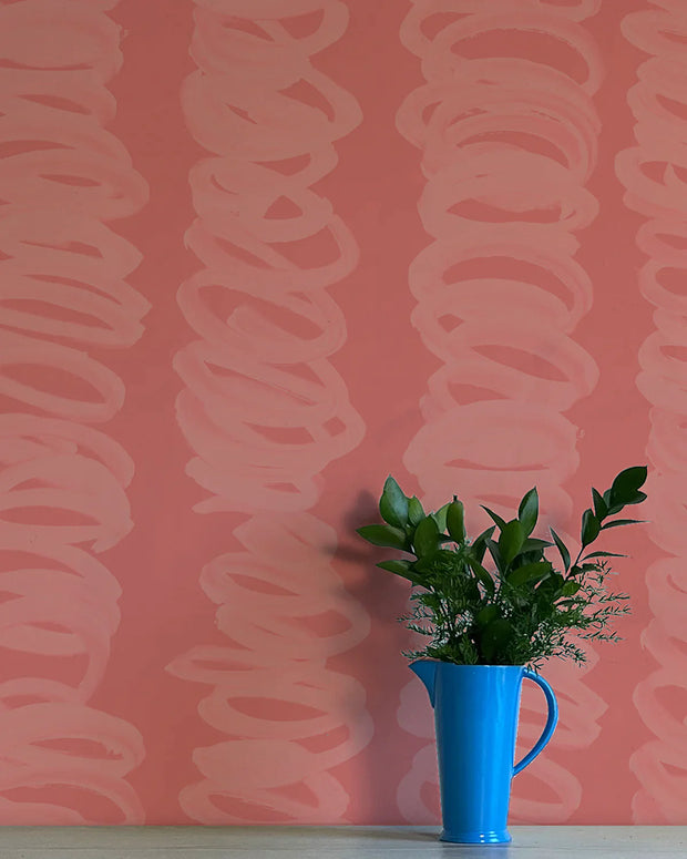 A section of pink wallpaper with a contemporary abstract pattern, next to a blue vase with green leaves.