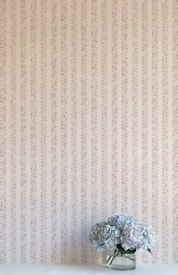 A wall adorned with pink wallpaper featuring a dotted pattern of slipper shells, flanked by a vase with purple flowers and a clear glass object.