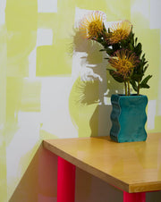 Teal vase with yellow flowers on a wooden table against a colorful wall. Electric Sunshine on White Wallpaper – Agolise | Half-Drop 10 Yard Roll