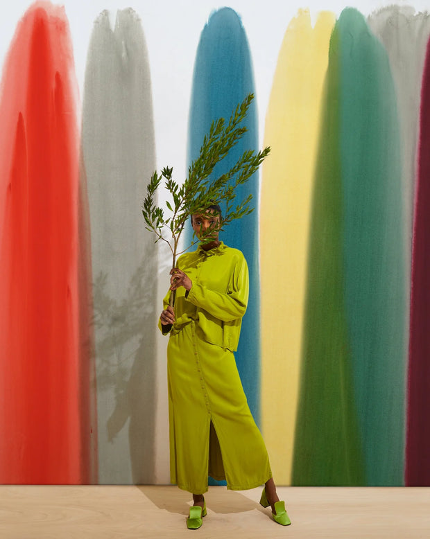 Person in a bright green outfit holding branches against colorful surfboards. Prism Wallpaper – Flood Mural | Custom Type II PVC Free