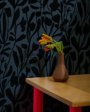 Brown vase with flowers on a wooden table against a dark floral-patterned wall.