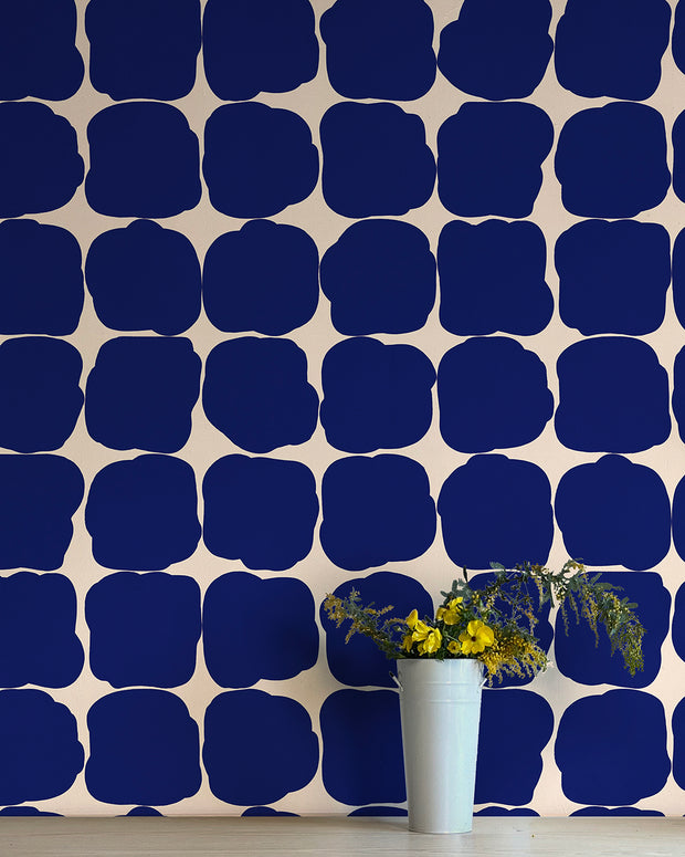 A wall adorned with blue geometric patterned wallpaper, featuring a vase with flowers in the foreground.  Blueberry Wallpaper by Blot | Non Woven Double Roll, 27” Trimmed