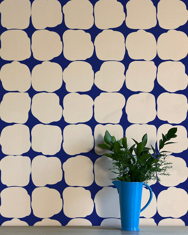 A section of wallpaper with a geometric pattern featuring beige, blue, and white colors, shown with a blue mug and green plant on a table against the same wall. Cold Cream Wallpaper by Blot | Non Woven Double Roll, 27” Trimmed