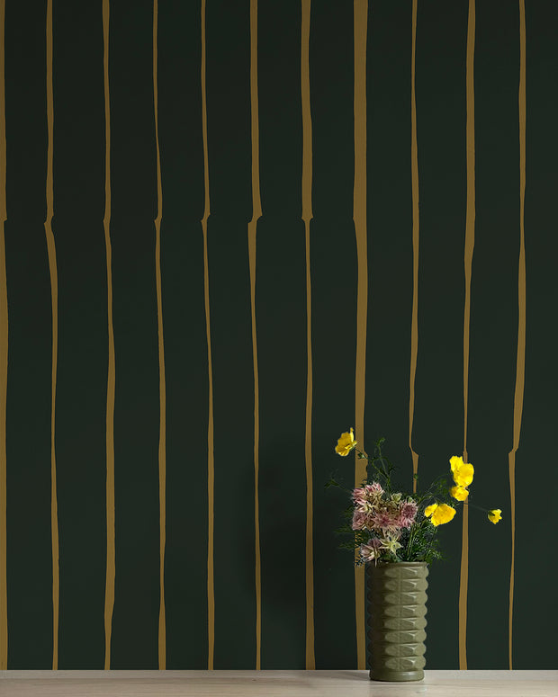 A wall adorned with green and yellow stripes wallpaper, flanked by a vase with flowers and a decorative object in the background. Arles Wallpaper – Lane | Non Woven Double Roll Design