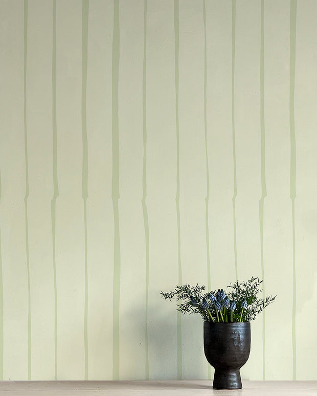 A wall adorned with green and white striped wallpaper, featuring a vase with blue flowers on the floor. Cucumber Wallpaper – Lane | Non Woven Double Roll Design