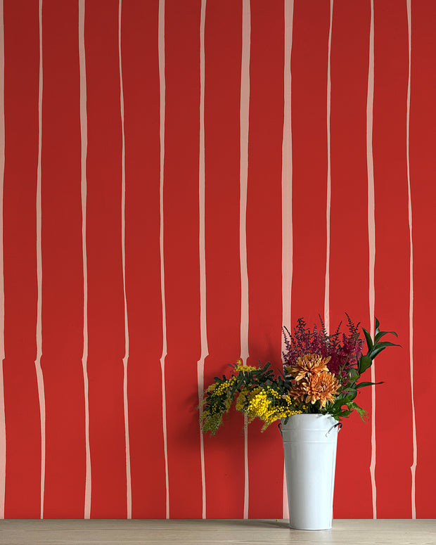 A wall adorned with red wallpaper featuring white vertical stripes, flanked by a white vase containing various flowers. Toulouse Wallpaper – Lane | Non Woven Double Roll Design