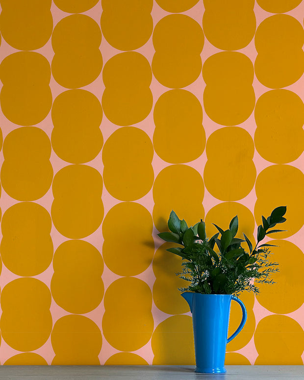 A wall adorned with yellow and pink geometric patterned wallpaper, featuring a blue vase with green leaves on a table. Barbara Wallpaper – Venn | Non Woven Double Roll Straight Match
