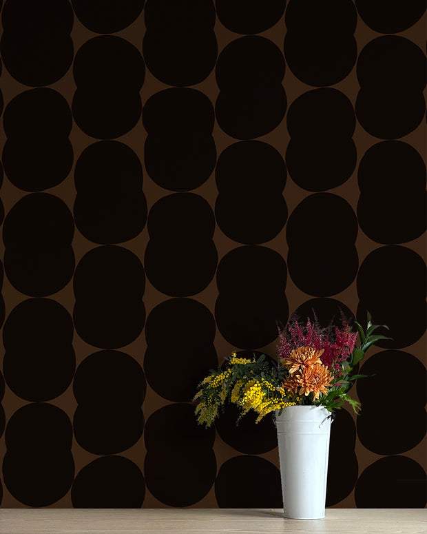 A vase with flowers placed on a surface with a black, geometric patterned wallpaper. Bon Bon Wallpaper – Venn | Non Woven Double Roll Straight Match