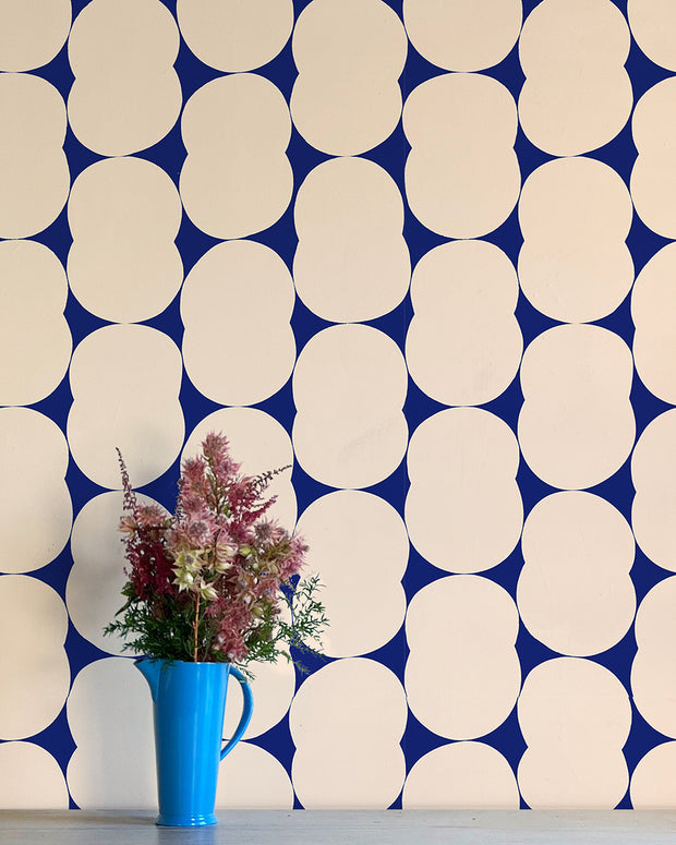 A section of wallpaper with a geometric pattern featuring circles in various shades of blue and beige, with a blue mug and pink flowers positioned against the wall.  Cosmic Wallpaper – Venn | Non Woven Double Roll Straight Match