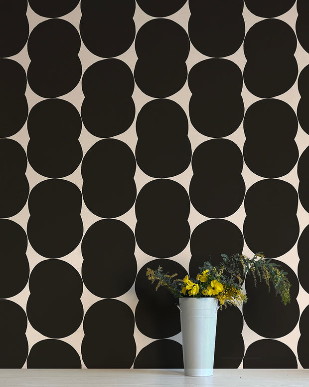 A wall adorned with black geometric shaped wallpaper, featuring a pattern of circles, and a white vase with yellow flowers and green leaves on a table in the foreground. Shapeshift Wallpaper – Venn | Non Woven Double Roll Straight Match