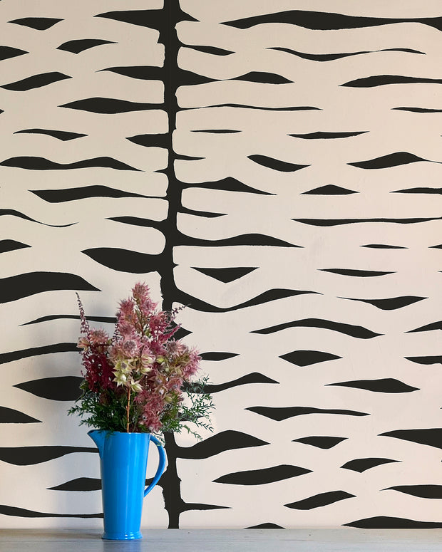 A wall adorned with a geometric pattern of black and white zigzag lines, featuring a blue vase with pink flowers on a white surface. Bone White Wallpaper – Zag | Non Woven Double Roll Design