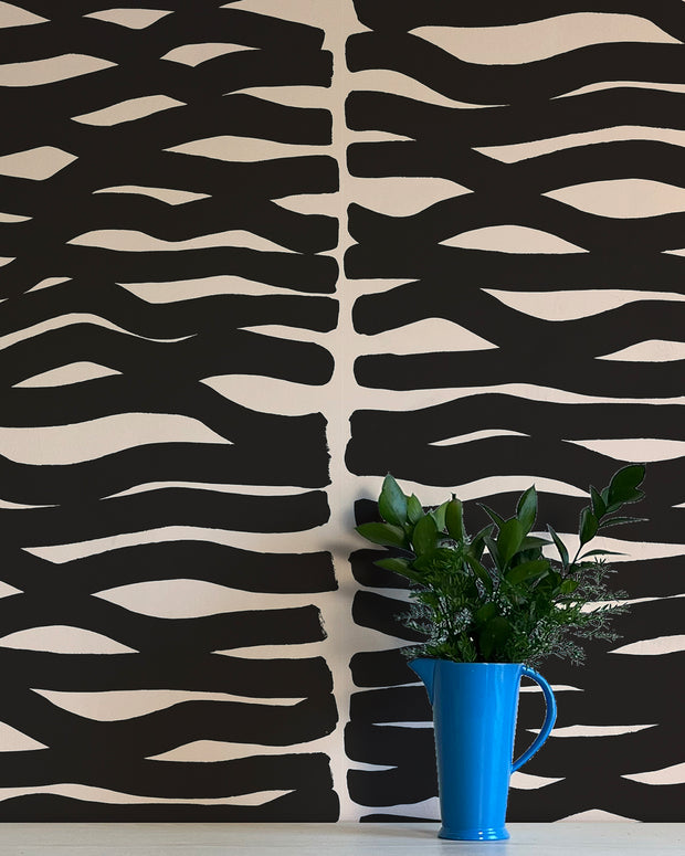 A wall adorned with black and beige geometric patterned wallpaper, featuring a blue vase with green leaves on a table in the foreground. Carbon Black Wallpaper – Zag | Non Woven Double Roll Design